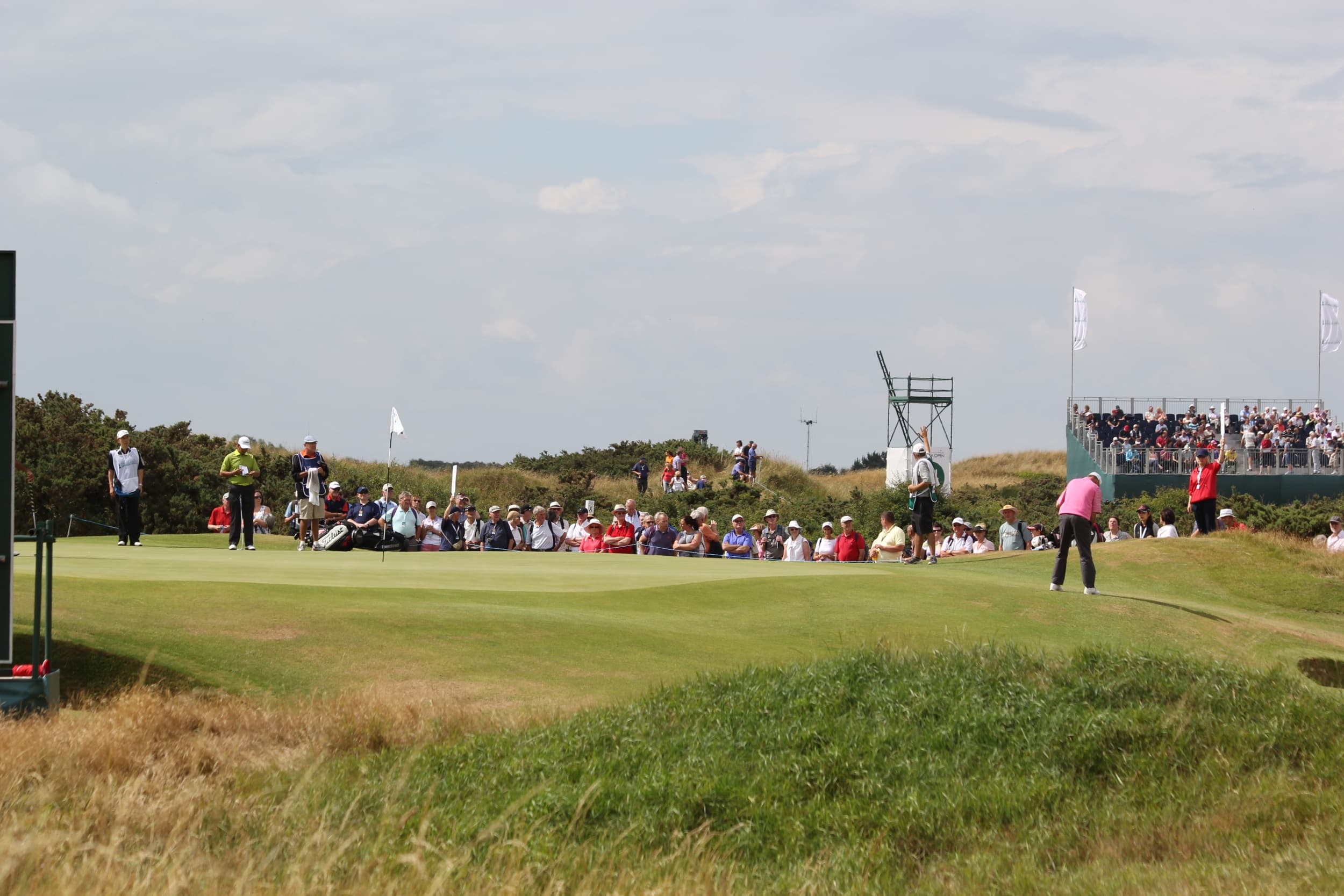 Packed grandstands lining the 14th green at Royal Birkdale during The Senior Open Championship, with links dunes and championship sky behind